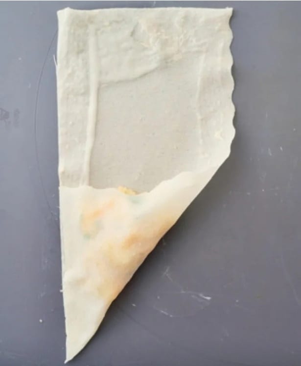 『三角春巻きの包み方』① 春巻きの皮(半分に切ったもの)を縦長に置き、皮の下半分の中央に具材をのせる。 手前以外の 3 辺にのりをぬる。具材がはみ出さないよう、手前の辺を左の辺に重ねるように折り、重なった辺どうしを指で押さえて密着させる。