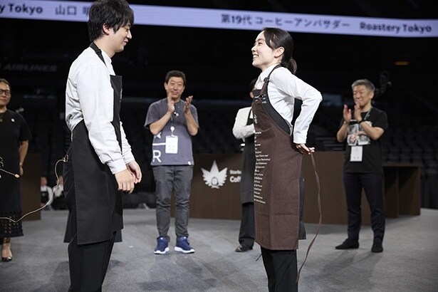 第18代コーヒー アンバサダーの下出さんから山口さんへ、歴代のコーヒーアンバサダーの名前が刺繍されたコーヒー染めのエプロンが贈呈された。エプロンには新たに彼女の名前が刻まれる