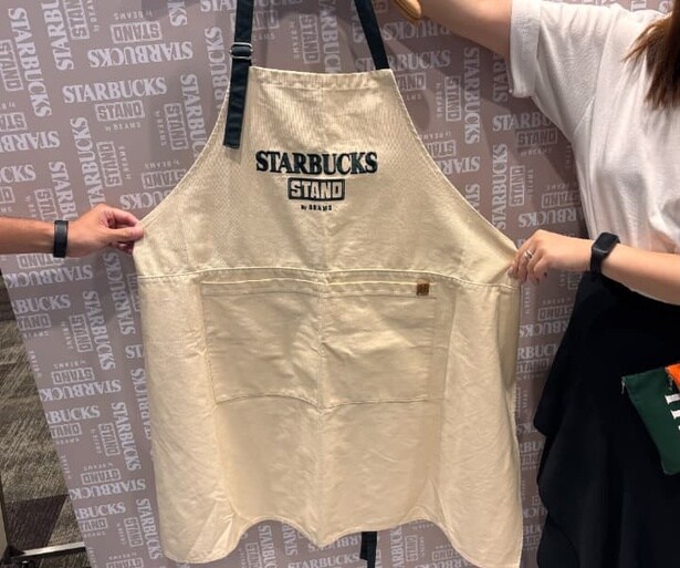 胸元に「STARBUCKS STAND by BEAMS」のロゴを大きく刺しゅうであしらったエプロン。オフホワイトとブラウンの2色で展開
