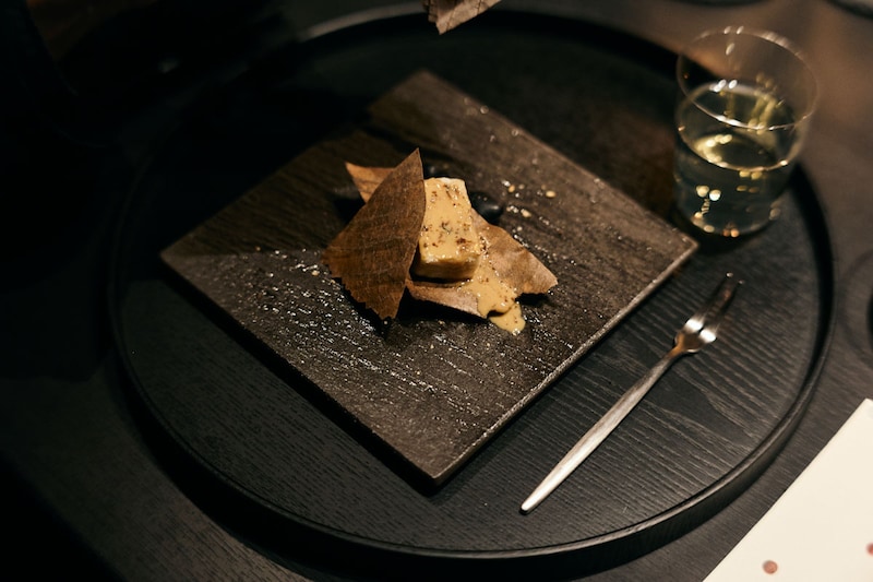 「聴く」テーマにした一皿。炭火の香りをまとったごま豆腐。皿の下に敷かれた玉砂利が擦れ合う音は、余市蒸溜所で守り続けられてきた石炭直火蒸溜の音と重なります[食楽web]