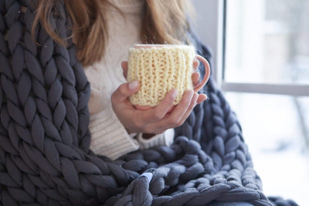 ブランケットを肩にかけて窓辺で温かい飲み物を飲む女性