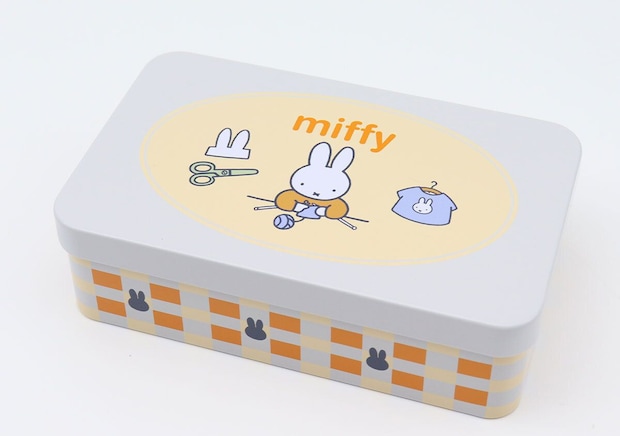 白い長方形の缶ケースのふた、中央の黄色い楕円フレーム内にハサミと編み物するミッフィーとミッフィーTシャツが描かれ、側面はオレンジと水色のチェック柄