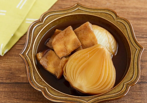 茶色い器に盛った新玉ねぎと油揚げのスープ煮 半分に切った新玉ねぎと四角く切った油揚げを、濃い茶色のだし汁で煮たスープ煮を、茶色い花型の器に盛った写真