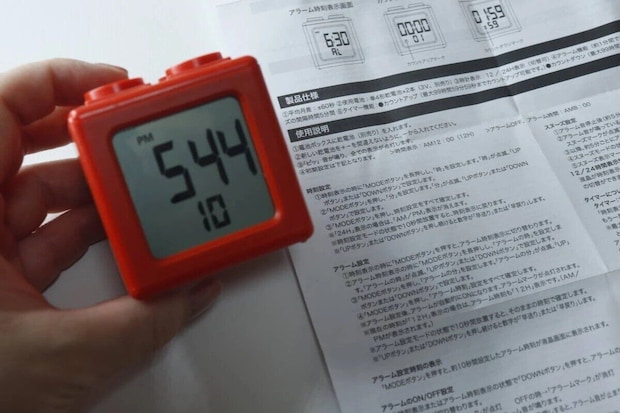 赤いブロック型デジタルタイマー時計を手に持ち、時刻表示「5:44」が見える。横には説明書
