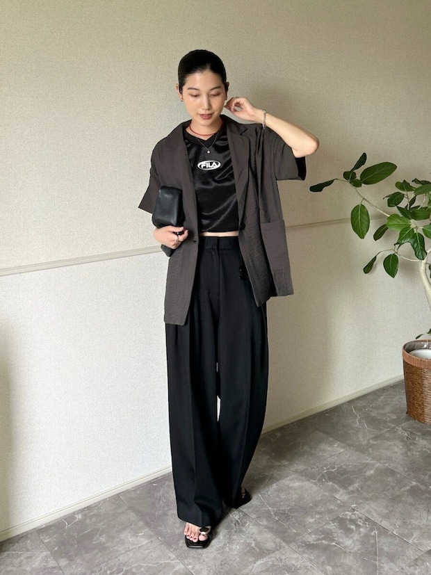 濃いグレーの半袖ジャケットと黒のロゴトップス、黒ワイドパンツを着た女性。 濃いグレーの半袖ジャケットと黒のロゴトップス、黒ワイドパンツを着た女性。