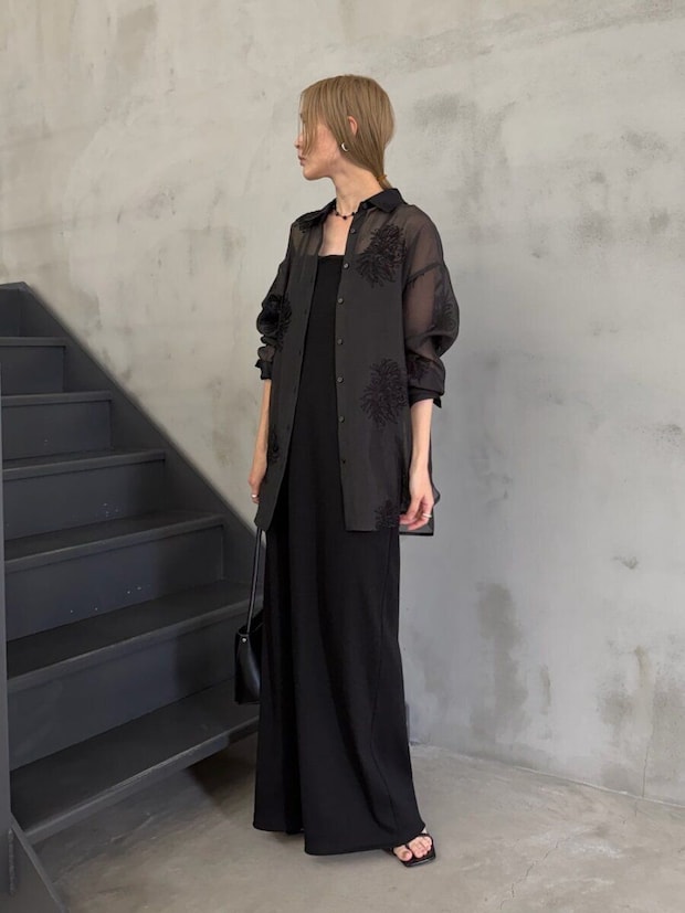 黒い刺繍入りシアーシャツと黒いキャミワンピースを着用し、階段の横に立つ女性