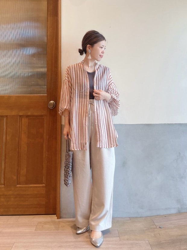 ストライプ柄シアーシャツとベージュのワイドパンツを着用し、バッグを持つ女性