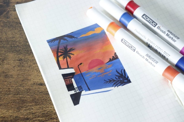 方眼紙にアクリルマーカーで描かれた夕焼けの風景画と、その横にマーカー3本が置かれている