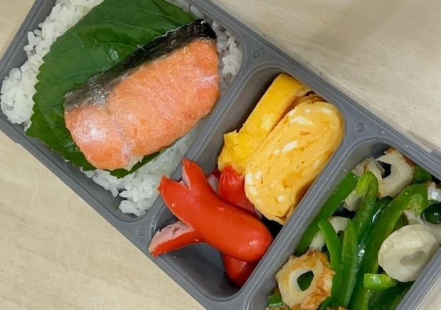 グレーの薄型弁当箱に、ご飯、焼き鮭、卵焼き、ウインナー、ピーマンとちくわの炒め物などのおかずが詰められている