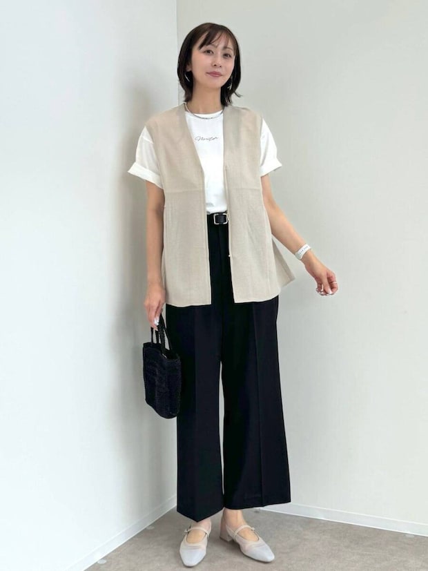 ベージュジレと白T、黒パンツを着た女性が壁を背景に立つ ベージュジレと白T、黒パンツを着た女性が壁を背景に立つ
