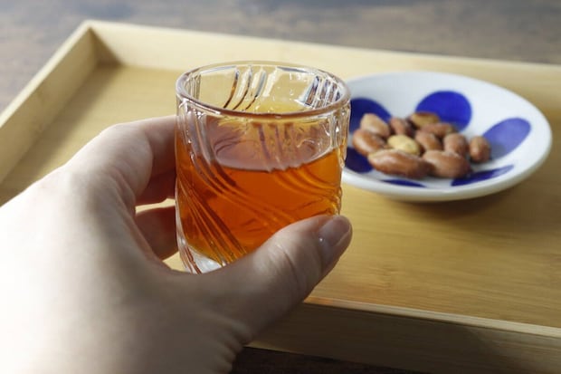 ミニグラスに入った琥珀色の飲み物を持ち、奥におつまみ ミニグラスに入った琥珀色の飲み物を持ち、奥におつまみ