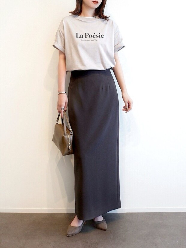 ベージュのロゴTシャツと濃いグレーのタイトスカートを着た女性が茶色のバッグを持っている