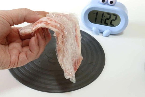 女性の手がラップされた冷凍肉を持ち、黒い解凍プレートと時計が写っている