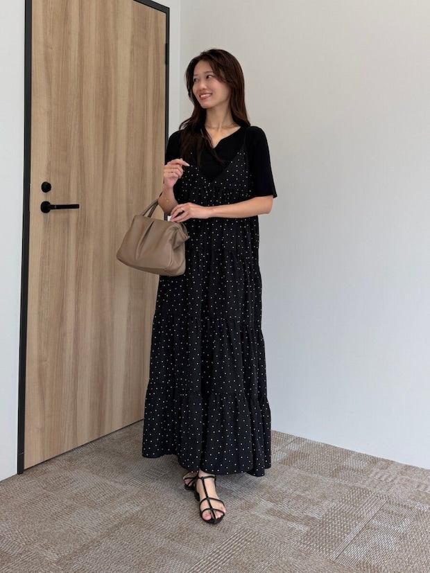 ドット柄の黒キャミワンピに黒ニットを重ね、ベージュのバッグを持つ女性が笑顔で立っている ドット柄の黒キャミワンピに黒ニットを重ね、ベージュのバッグを持つ女性が笑顔で立っている