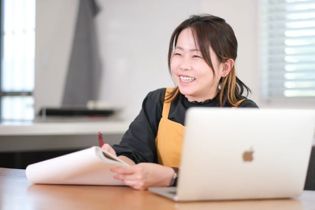 黄色いエプロンの女性が笑顔でノートにペンを走らせている