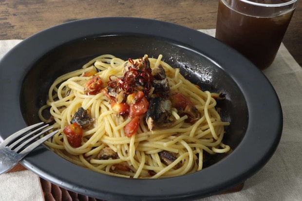 黒い皿に盛り付けられたいわしとミニトマトのオイルパスタ。奥にはグラスに入った飲み物が見える
