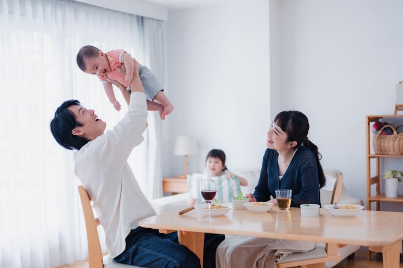 リビングに、夫婦（男女）と乳児、幼児がいる。父親と見られる男性が乳児を「たかいたかい」している