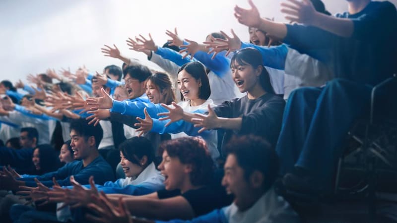 観客席のような場所で、多くの人々が両手を前に伸ばし、力強く動かしている。「サインエール」を送っている様子。