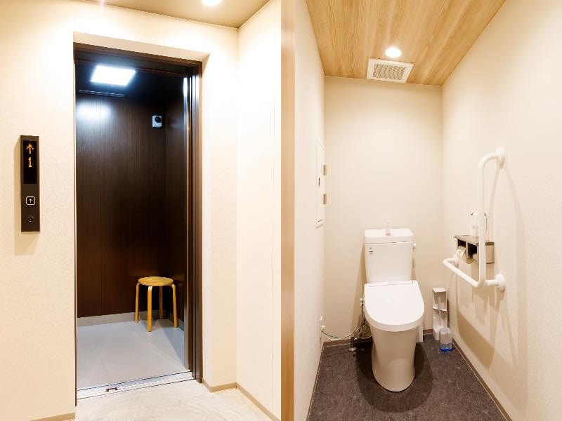 施設内のエレベーターホールとトイレの写真。エレベーターの内部には小さな丸椅子がある。トイレには、手すりが設置。