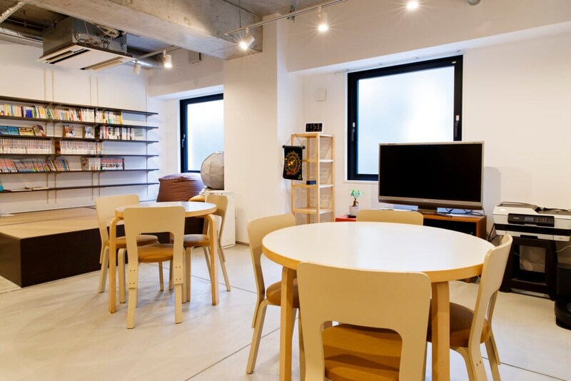 共有スペースの室内写真。左奥にはマンガや本が並べられた壁一面の棚があり、その前には座布団のようなものと畳スペース。部屋の中央には、木製の丸いテーブルが2つ、それぞれ椅子が4脚ずつ配置。右奥には、大きなテレビとプリンター、棚がある。窓は2つあり、すりガラスのようになっている。