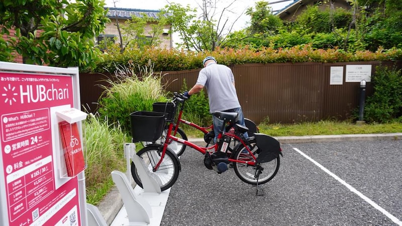 「HUBchari」と書かれた赤い看板の前に、赤と黒の電動アシスト自転車が数台駐輪されている。一人の男性が自転車を動かそうとしている様子。