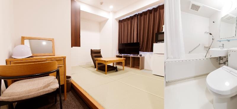 左：居室 
和室の畳の上に低いテーブルと座椅子、テレビと小型冷蔵庫が設置。奥の窓には濃い茶色の遮光カーテンがかかっている。手前には鏡付きのドレッサーと木製の椅子、白いヘルメットが置かれている。

右：浴室 白を基調とした清潔な浴室で、浴槽と洋式トイレ、洗面台が一体となったユニットバス。